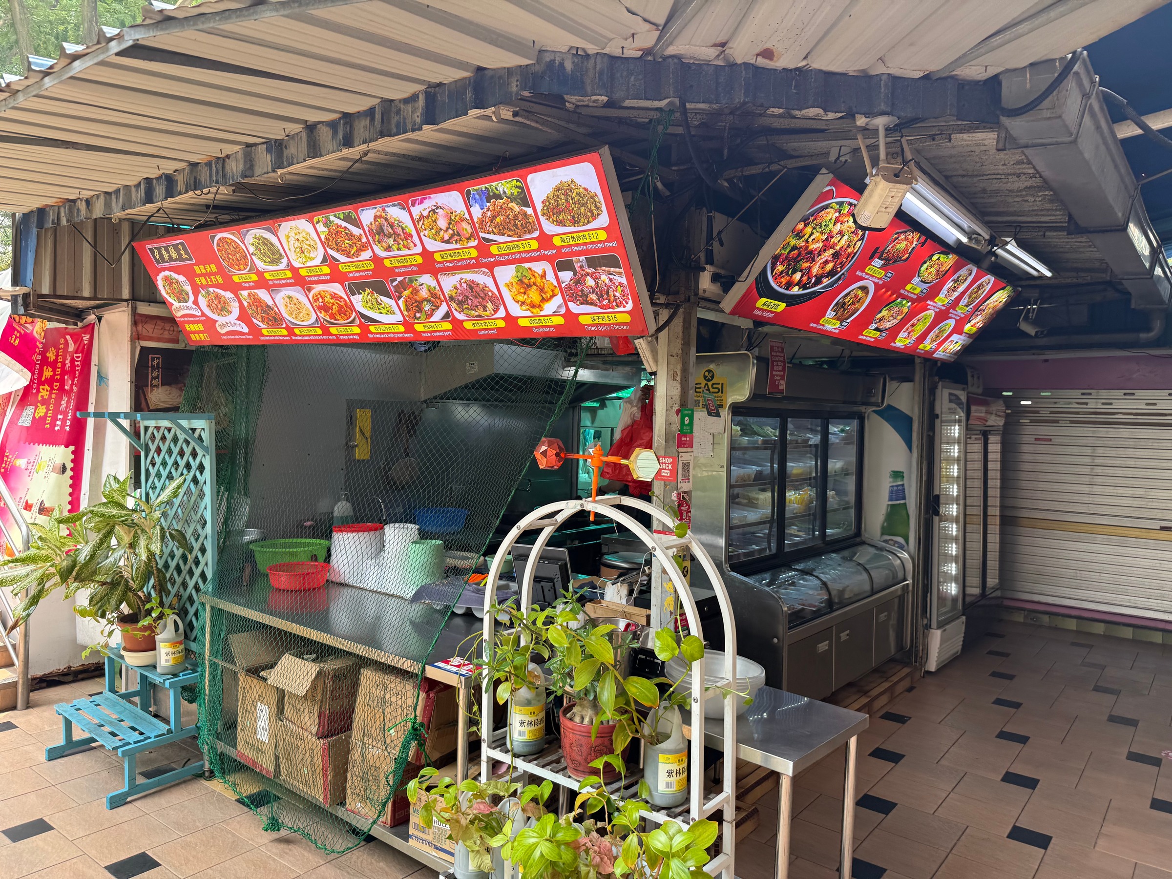 An outdoor food stall under an awning, surrounded by potted plants.