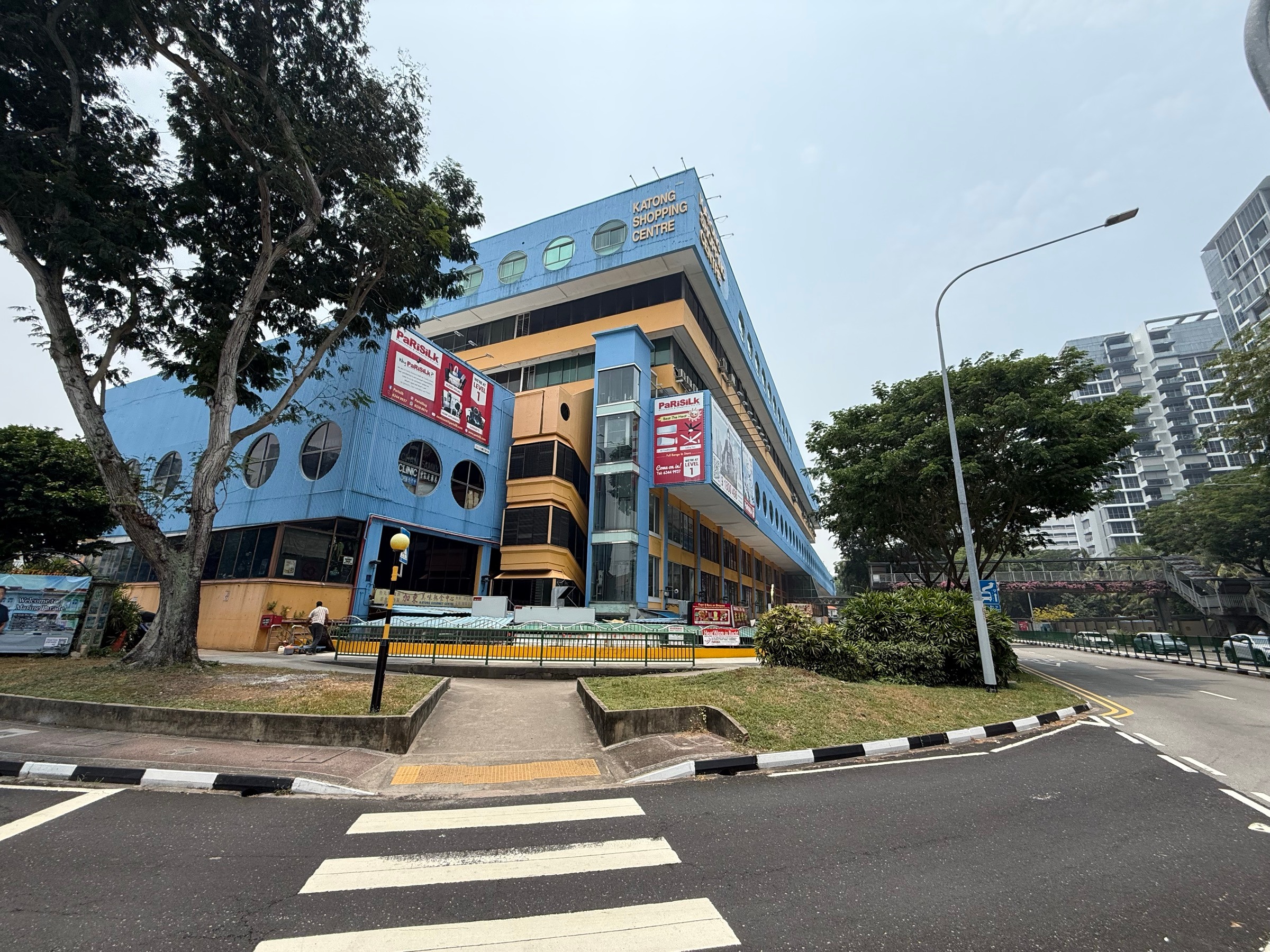 The blue-and-yellow end of Katong Shopping Centre beside the road junction, with round windows and a pedestrian crossing.