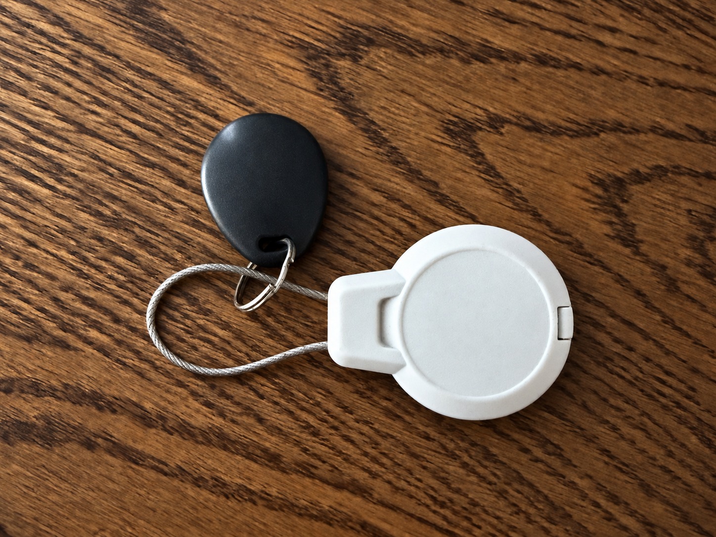 Black electronic access fob and white AirTag case joined by a steel cable on a wooden table