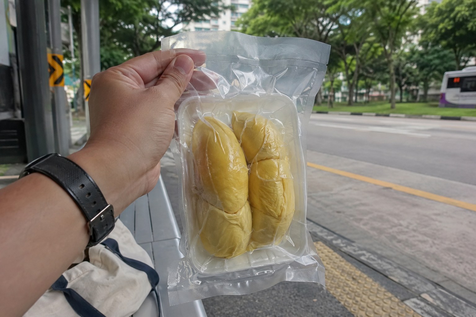 A hand holding vacuum-packed durian at a bus stop.