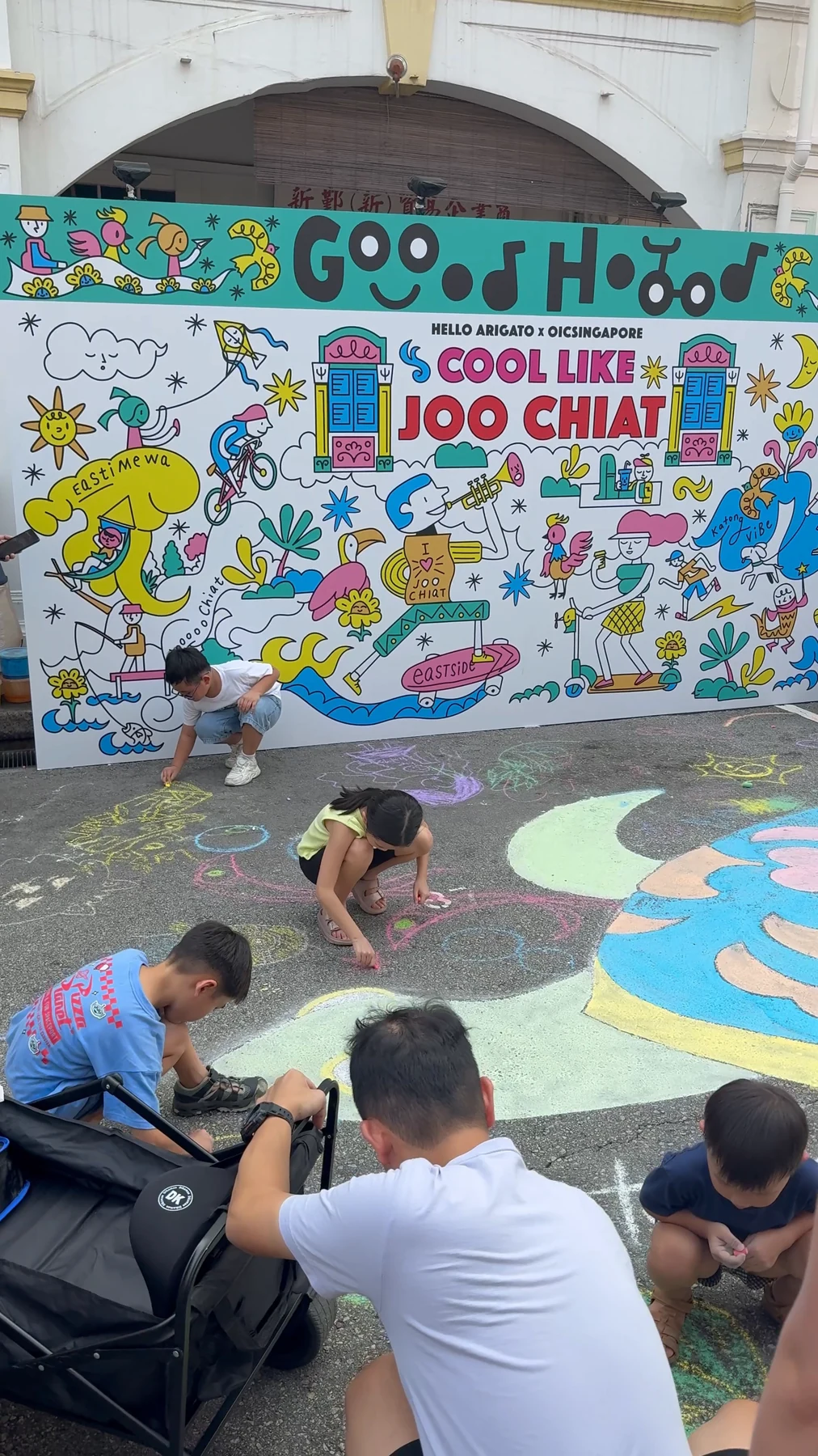 Children draw with coloured chalk on the road beside a bright wall.