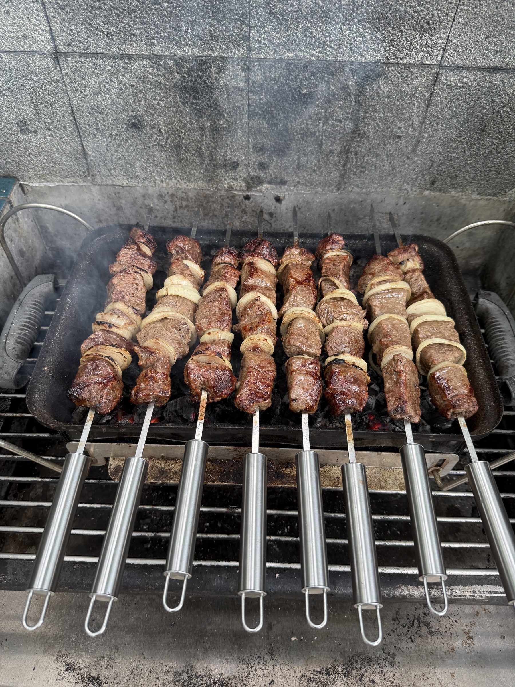 Browned shashlik and onion rings on flat skewers over charcoal.