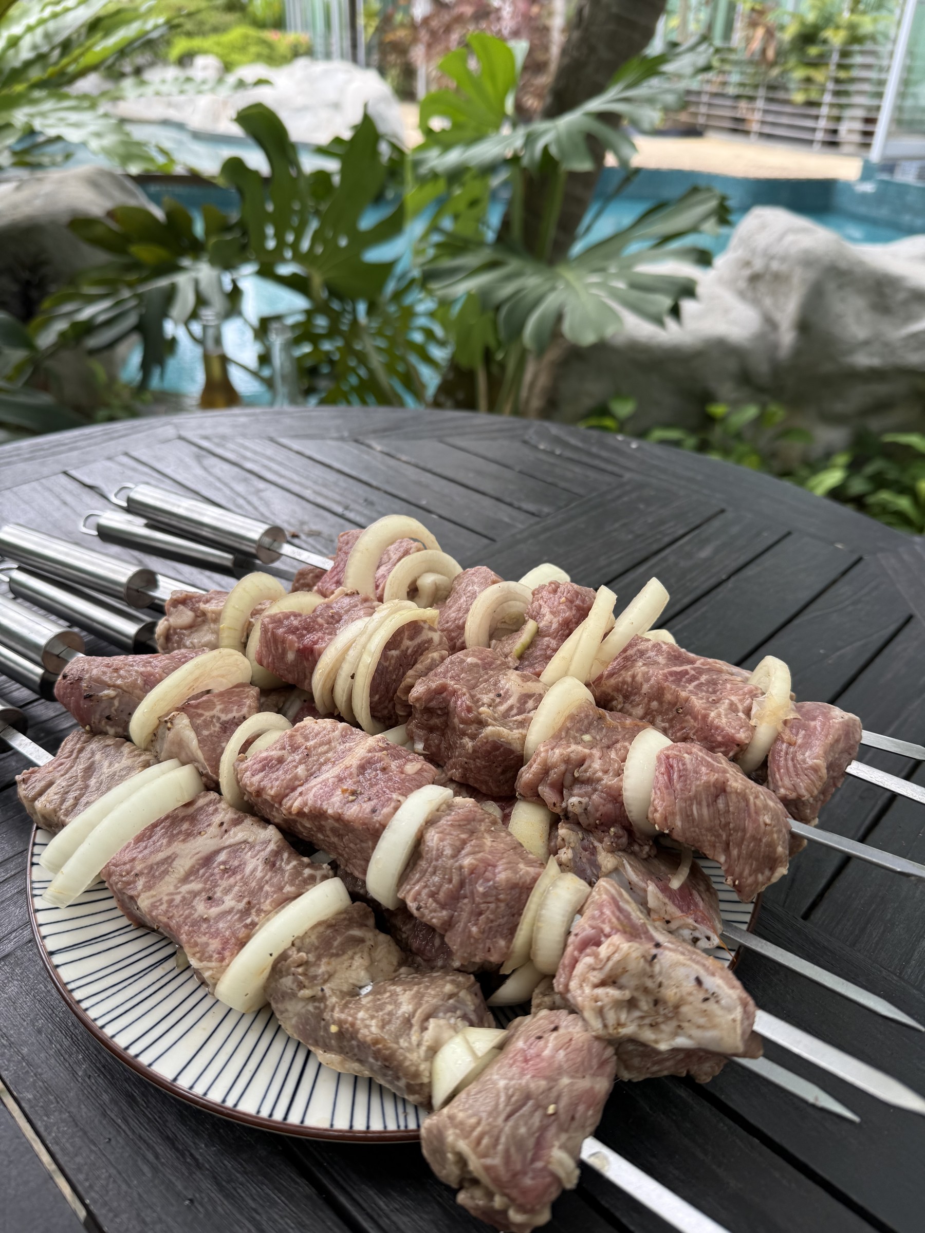 Marinated meat and onion rings threaded onto flat skewers beside a swimming pool.